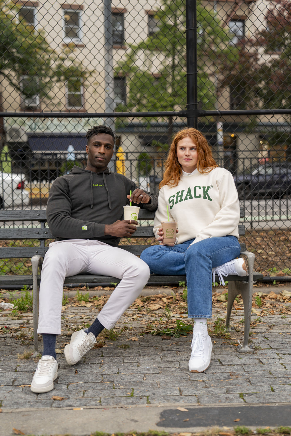 Two people sitting on a park bench wearing Shake Shack merch and enjoying Shake Shack menu items