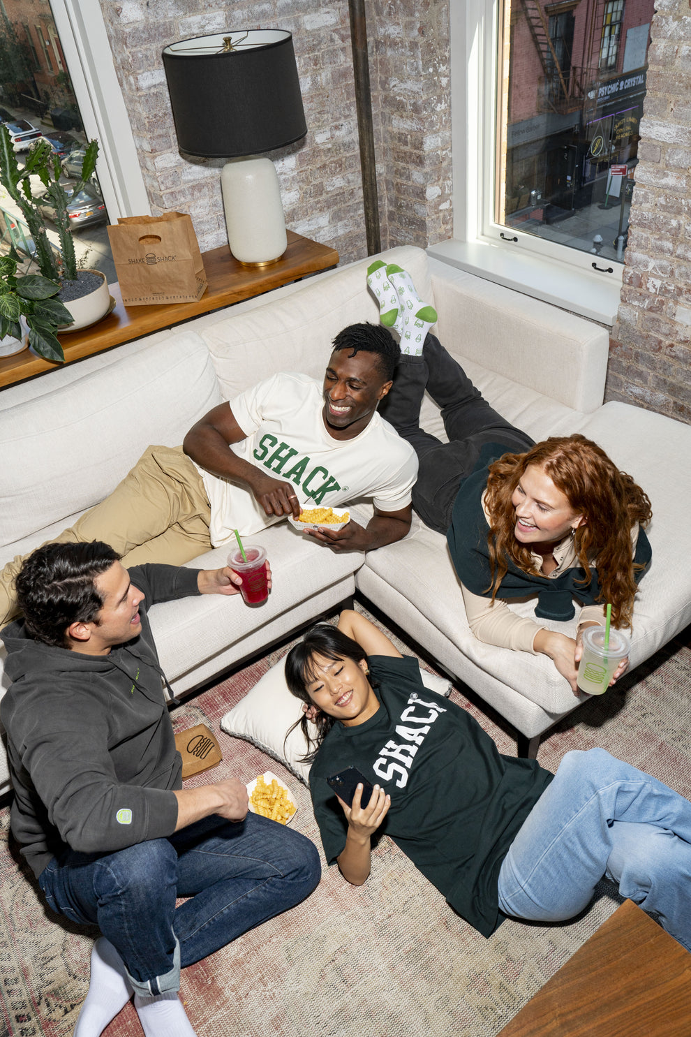 Four people sitting on or around a couch wearing Shake Shack merch and enjoying Shake Shack menu items