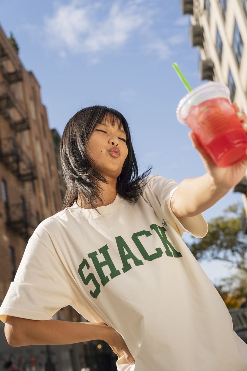 Girl outside wearing cream-colored Shake Shack tee and holding Shake Shack drink up to the camera