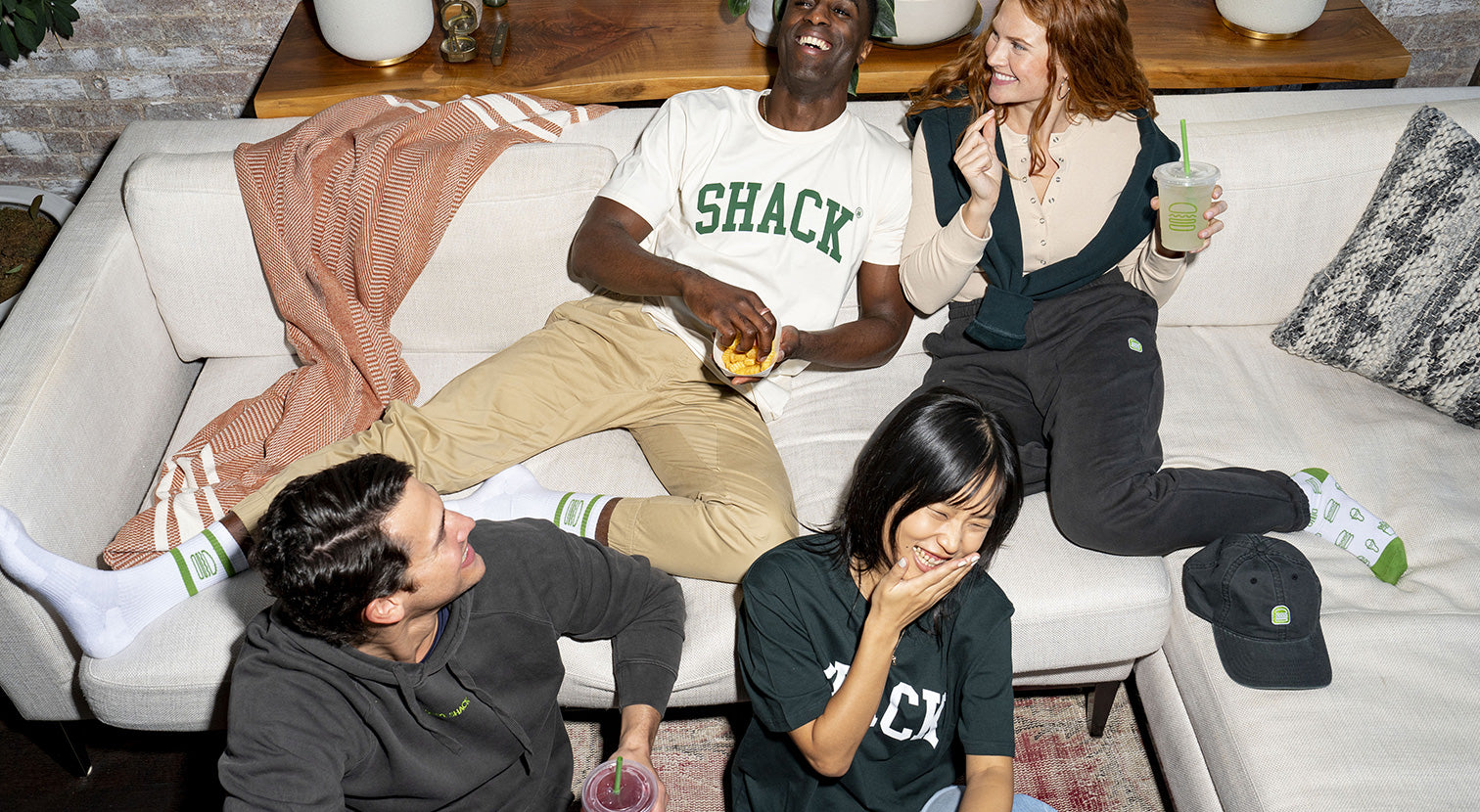 Four people sitting on or around a couch wearing Shake Shack merch and enjoying Shake Shack menu items